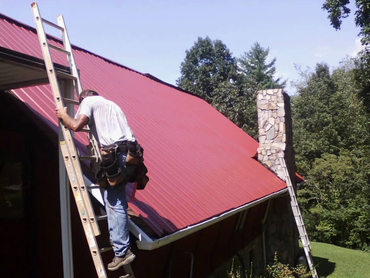 Roofer on a ladder beside a red metal roof and stone chimney — Soffit & Fascia Repair by Roofing Brothers in Cobalt